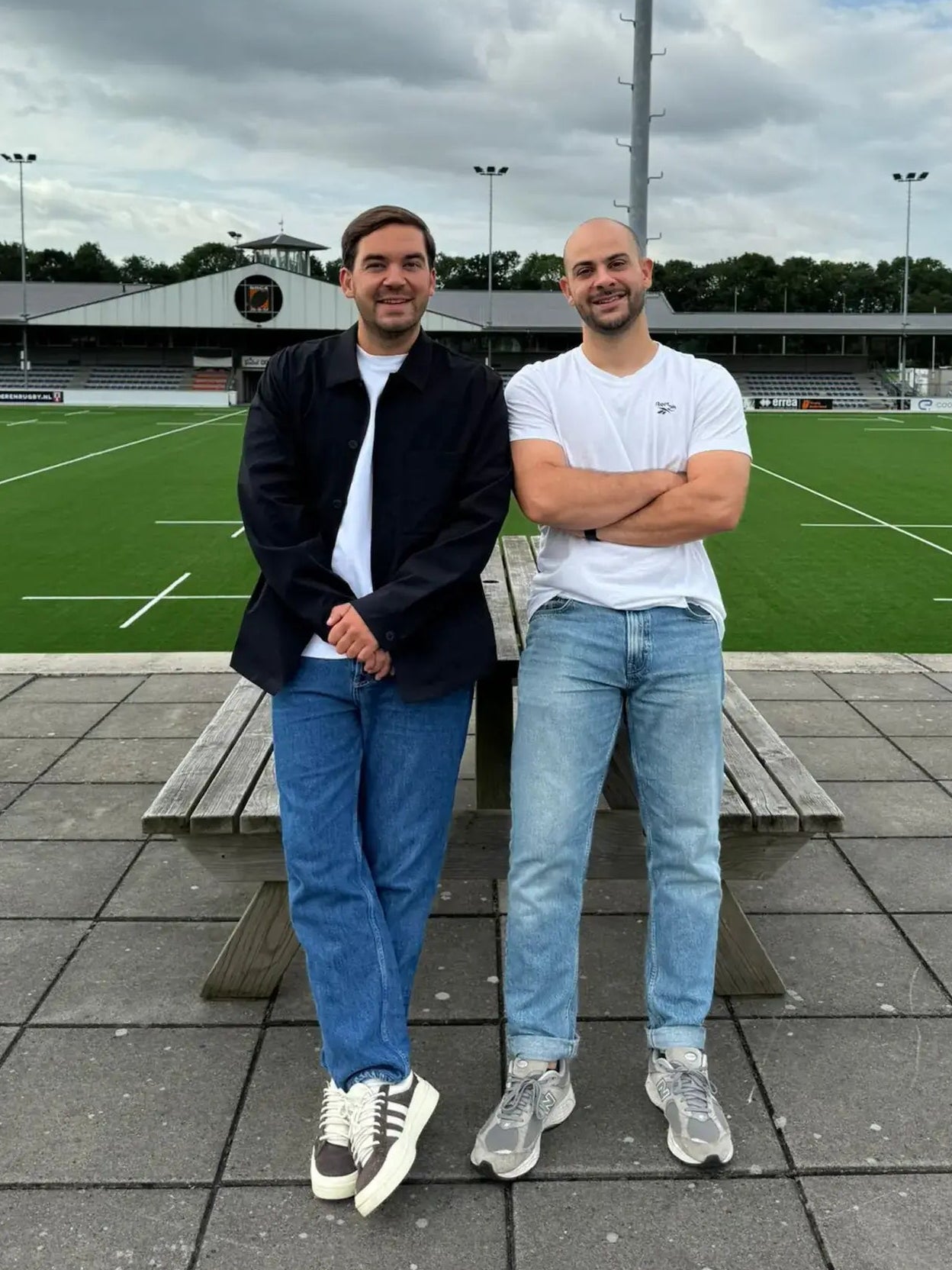 Brian en Nelson poserend in casual outfits (spijkerbroek, shirts en sneakers) op een picknicktafel bij een rugbyveld.